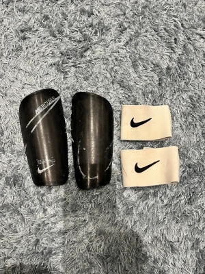 Nike Mercurial benskydd med vita straps - Säljer ett par svarta Nike Mercurial benskydd med tillhörande vita elastiska straps med svart Nike-logga. 