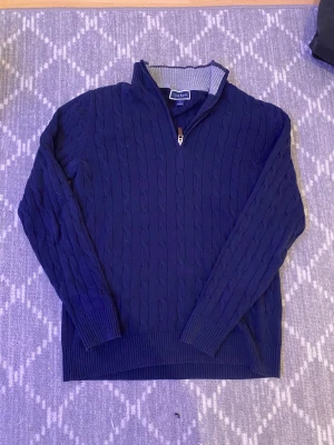 Marinblå stickad tröja Club Room  - Snygg marinblå stickad tröja från Club Room i storlek S. Tröjan har kabelstickat mönster, ribbade muddar och krage med grå insida. Half zip-dragkedja framtill med läderdetalj. Perfekt för dig som gillar klassisk stil och vill ha något varmt och bekvämt.