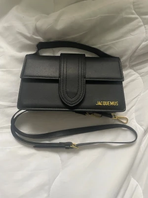 Svart Jacquemus handväska i skinn - Snygg svart handväska från Jacquemus i stilren, rektangulär form med guldfärgade detaljer och logga. Väskan har både handtag och avtagbar axelrem med guldfärgade spännen. Tillverkad i slätt skinn och har en elegant design som sticker ut.