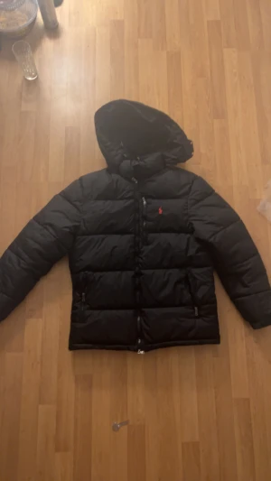 Svart pufferjacka Polo Ralph Lauren - Svart pufferjacka från Polo Ralph Lauren med huva och två sidofickor med dragkedja. Jackan har det klassiska röda Polo-logot broderat på bröstet och quiltad design. Perfekt för kalla dagar och har en snygg, clean look.