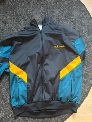 Adidas svart och blå vindjacka - Snygg vindjacka från Adidas i svart med blå och gula detaljer. Jackan har dragkedja framtill, ribbade muddar och två sidofickor. Adidas-logga i gult på bröstet. Perfekt för dig som gillar sportig stil och färgkombinationer.