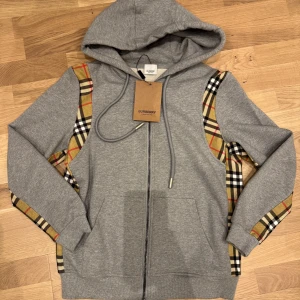 Grå Burberry hoodie - ny med tagg! - Snygg grå hoodie från Burberry med klassiska rutiga paneler längs ärmarna. Tröjan har dragkedja framtill, huva med snörning och två fickor. Perfekt för dig som vill ha en stilren streetlook med lyxig touch.