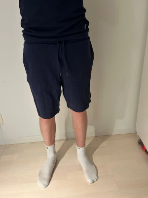 Marinblå shorts från Lyle & Scott - Säljer ett par marinblå shorts från Lyle & Scott med klassisk gul logga på benet. De har snörning i midjan, sidofickor och en bakficka. Materialet är mjuk bomull och passformen är loose, perfekt för chill dagar. Enkla och stilrena med sportig vibe.