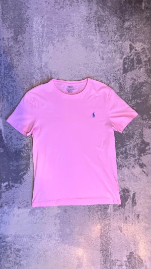 Rosa t-shirt från Polo Ralph Lauren - Snygg rosa t-shirt från Polo Ralph Lauren med klassisk rund hals och korta ärmar. T-shirten har en liten blå Polo-logga broderad på bröstet och är gjord i mjuk bomull som känns skön mot huden. Perfekt för dig som gillar stilrena plagg med en touch av färg.