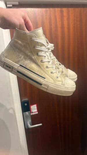 Dior skor - Säljer ett par high-top sneakers från Dior med ikoniskt monogrammönster. Skorna har snörning, rund tå och platt sula. Snygga detaljer med Dior-logga på sidan och genomskinliga paneler som ger en modern vibe. Skriv om ni vill ha fler bilder.