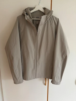 Beige vindjacka från Uniqlo med huva - Snygg beige vindjacka från Uniqlo med huva och dragkedja framtill. Jackan är vattenavvisande, vindtät och har ett stilrent, minimalistiskt utseende. Perfekt för dig som gillar clean look och funktionella plagg. Passar nog även en mindre large.