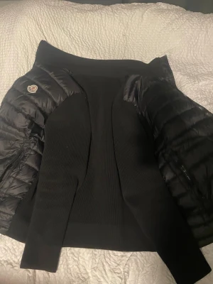 Svart Moncler hybridjacka - Säljer en svart Moncler hybridjacka med quiltad front i dun och stickade ärmar. Jackan har dragkedja, hög krage och två fickor fram. Klassisk Moncler-logga på bröstet och coola detaljer på insidan. Perfekt för dig som vill ha en clean och stilren look. M-L