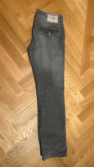 Grå jeans från True Religion - Snygga grå jeans från True Religion med klassisk logga på bakfickan och kontrastsömmar. Modellen har raka ben och en cool fickdetalj med lock och vit söm. Perfekt för dig som gillar streetwear och vill sticka ut med ett ikoniskt märke. Pris kan diskuteras vid en snabb affär