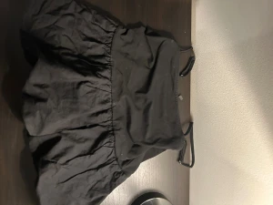 Svart kort topp från Divided - Supersöt svart topp från Divided med tunna axelband och voluminös kjol. Enkel och stilren design som ger en trendig look. Perfekt för dig som gillar minimalistiska plagg med lite extra volym. Materialet känns lätt och bekvämt.