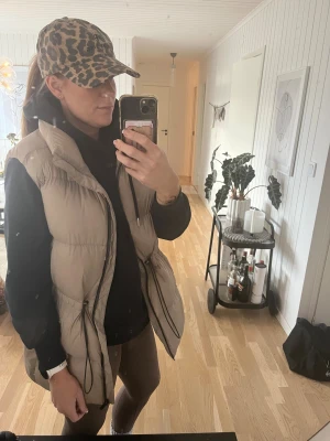 Beige dunväst från Stronger - Säljer en beige dunväst från Stronger med hög krage och svarta dragskor i midjan. Västen har quiltad design och dragkedja framtill. Perfekt att slänga över en hoodie för en chill look.