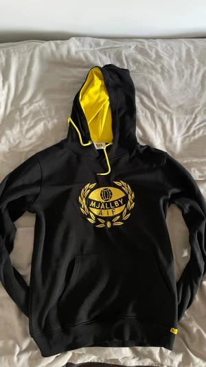 Svart och gul Mjällby AIF hoodie - Snygg svart hoodie från Mjällby AIF med gul insida på huvan och gul logga framtill. Tröjan har snörning vid huvan, stor magficka och ribbade muddar. Perfekt för dig som vill visa ditt stöd för laget och gillar sportig stil.