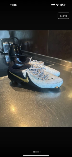 Nike fotbollsskor i svart och vitt - Snygga fotbollsskor från Nike med svart och vit ovandel, guldigt swoosh på sidan och snörning. Skorna har platt sula med dobbar för bra grepp på planen. Materialet är syntet och designen är sportig med rund tå.