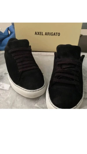 Svarta sneakers från Axel Arigato - Snygga svarta sneakers från Axel Arigato med ovandel i mocka och detaljer i skinn. Skorna har vit platt sula och klassisk snörning. Perfekta för dig som gillar stilrena och minimalistiska sneakers med premiumkänsla. Funkar svin bra till stilen grisch osv, du får även med boxen