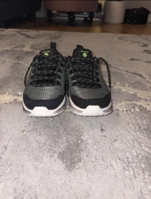 Svarta Skechers sneakers med gröna detaljer - Säljer ett par svarta Skechers sneakers från serien 'Luminators' med gröna detaljer och reflekterande mönster. Skorna har snörning, vit platt sula och är tillverkade i syntetmaterial. De har USB-port för laddning och coola lysande effekter – perfekt för dig som vill sticka ut.