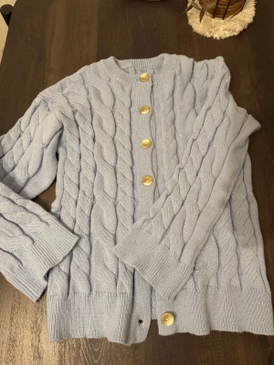 Ljusblå stickad kofta med knappar - Säljer en ljusblå stickad kofta med grova flätstickade detaljer och rund hals. Koftan har stora, ljusa knappar framtill och ribbade muddar vid ärmslut och nederkant. Perfekt för dig som gillar mysiga och stilrena plagg.
