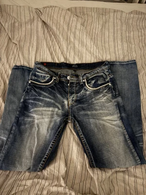 Blå jeans från Smog med kontrastsömmar - Snygga blå jeans från Smog med slitna detaljer och kontrastsömmar. Klassisk femficksmodell med vita och gula sömmar, samt en cool patch bak i midjan. Jeansen har en rak passform och är tillverkade i jeansmaterial.