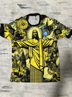 Nike Brasil CBF gul grafisk t-shirt - Unik gul och svart Brasil CBF t-shirt från Nike med ett stort grafiskt tryck av Kristusstatyn och brasilianska motiv. T-shirten har korta ärmar, rund hals och broderade detaljer med CBF-emblem och Nike-logga. Perfekt för dig som gillar bold streetwear och fotbollsinspirerad stil.