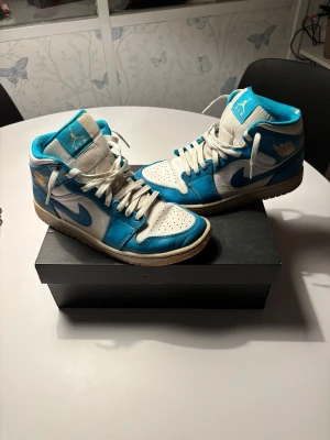 Nike Air Jordan 1 UNC blå och vita - Säljer ett par Nike Air Jordan 1 i den ikoniska UNC-färgen. Skorna har en snygg kombination av blått och vitt skinn, klassisk snörning och hög siluett. Perfekta för dig som vill sticka ut med en retro basketstil. Swoosh-logga på sidorna och Air Jordan-märke vid ankeln.