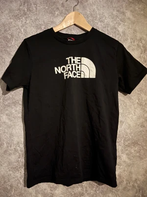 Svart t-shirt från The North Face  - Svart t-shirt från The North Face i storlek XL Youth/Junior. Klassisk rund hals och korta ärmar. Stor vit logga framtill och mindre logga på ryggen. Tillverkad i mjuk bomull som känns skön mot huden. Perfekt för dig som gillar streetwear och enkel stil.