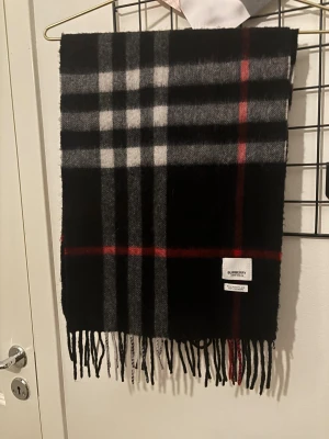 Rutig svart halsduk från Burberry - Snygg halsduk från Burberry i klassiskt rutigt mönster med svarta, gråa, vita och röda toner. Gjord i 100% kashmir. Mjuka fransar längs kanterna och stilren design som lyfter din outfit.