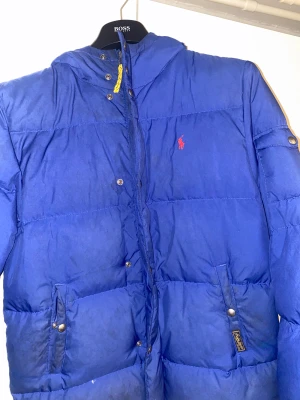 Blå pufferjacka från Polo Ralph Lauren - Säljer en blå pufferjacka från Polo Ralph Lauren med röd logga på bröstet och röd insida. Jackan har huva, dragkedja och tryckknappar framtill samt två fickor med knappstängning. Perfekt för kalla dagar och har en schysst streetstil.