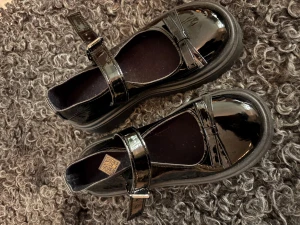 Svarta lackade ballerina med rem - Snygga svarta ballerina från Zara i glansigt lackmaterial med bred rem över foten och chunky sula. Skorna har rund tå och en cool, modern vibe som passar till många outfits. Perfekta för dig som vill ha en klassisk men edgy look. Helt oanvända.