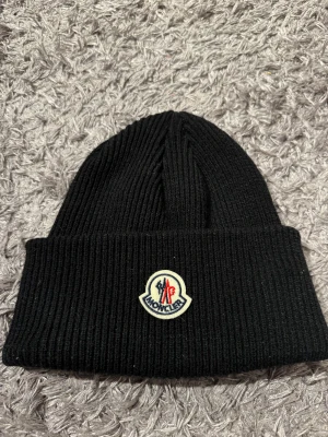 Svart ribbad Moncler mössa - Svart ribbstickad mössa från Moncler med klassisk uppvikt kant och broderad logotyp framtill. Snygg och enkel design som passar perfekt till kalla dagar. Materialet är mjukt och värmande, och märkesetiketten finns även på insidan.