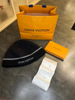 Louis Vuitton mössa - Snygg svart mössa från Louis Vuitton med grå kant och broderad logga framtill. Mössan har en klassisk form och är tillverkad i mjuk ull som håller dig varm. Perfekt accessoar för dig som vill ha en exklusiv och stilren look.