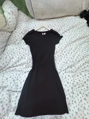 Svart kortklänning från H&M Divided - En svart, enkel och figurnära kortklänning från H&M Divided i storlek XS. Klänningen har rund halsringning och korta ärmar, tillverkad i mjuk och stretchig bomull. Perfekt för dig som gillar minimalistisk och stilren look.