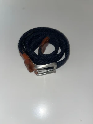 Mörkblå flätat bälte Jack & Jones - Snyggt mörkblått flätat bälte från Jack & Jones med bruna skinn-detaljer och en stilren silverfärgad metallspänne. Perfekt för att ge din outfit en schysst touch. Bältet har en klassisk design med tydlig branding på läderpartiet. Original pris: 250kr