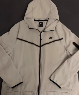 Nike Tech Fleece - Säljer en fräsch vit vindjacka från Nike med svart logga på bröstet och svarta detaljer längs dragkedjan och ärmsluten. Jackan har huva och hel dragkedja framtill, perfekt för chill dagar ute. Stilren design som passar till streetwear.