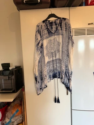 Blåvit blus från Cream med snörning - Säljer en flowy strandklänning från Cream i blått och vitt med paisleyinspirerat mönster och batikdetaljer. Blusen har v-ringning, långa ärmar och snörning med tofsar framtill. Materialet är lätt och luftigt, perfekt för varmare dagar. Storlek One size men passar kanske en L/XL. Mycket fint skick, jag hittar inga synliga anmärkningar eller defekter. 