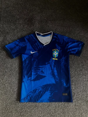 Brasil fotbollströja  - Snygg blå Brasilien fotbollströja från Nike med korta ärmar och V-ringad krage med vit kant. Tröjan har ett abstrakt mönster i olika blå nyanser och Brasilien-emblem med stjärnor på bröstet. Materialet är lätt och andas, perfekt för träning eller gym pga tajta passformen