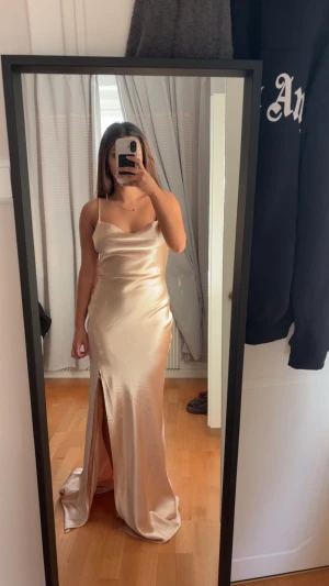 True decadence balklänning - Säljer en elegant långklänning i champagnefärgad satin med smala axelband, slits framtill och öppen rygg med knyt. Klänningen har en snygg draperad urringning och följsam passform som ger en lyxig känsla.