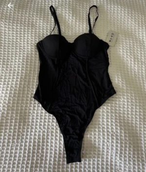 Svart body med bygel från NA-KD - Supersnygg svart body från NA-KD med bygel och formade kupor. Modellen har smala, justerbara axelband och hög skärning vid benen. Nypris är 400kr. Den har tyvärr inte prislappen  kvar men har ej använts. I storlek 70A