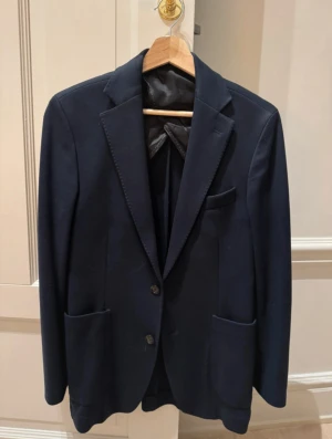 Oscar Jacobsson kavaj - Snygg mörkblå kavaj med två knappar framtill och klassisk krage. Kavajen har två stora fickor och en bröstficka, samt slät insida. Perfekt för dig som gillar stilrena och tidlösa plagg. Passar till både skjorta och t-shirt. Nypriset på kavajen ligger runt 3-4 tusen