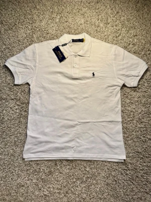 Vit Polo Ralph Lauren Piké - Klassisk vit pikétröja från Polo Ralph Lauren med ikonisk broderad logga på bröstet. Skjortan har korta ärmar, krage och knappar framtill. Tillverkad i mjuk bomull som andas och ger en clean look. Nypris ligger på 1499kr