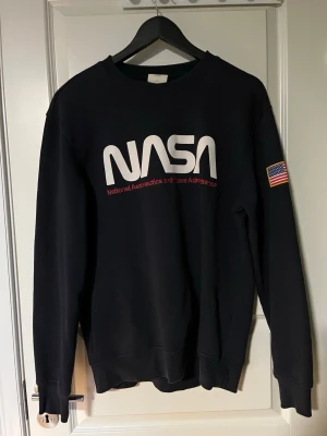 Marinblå pockettröja NASA H&M L.O.G.G. - Cool marinblå sweatshirt från H&M L.O.G.G. med stort NASA-tryck framtill och amerikansk flagga på soparmen. Klassisk rund hals och mjukt material, perfekt för dig som gillar rymdtema och streetwear.
