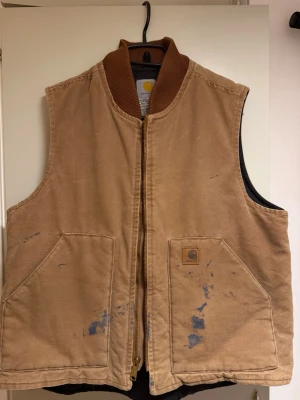 Beige Carhartt väst med dragkedja S/M - Klassisk beige väst från Carhartt med ribbad krage och två stora framfickor. Västen har en tydlig Carhartt-logga på fickan och stängs med dragkedja. Utsidan är i bomull och insidan har quiltad polyester. Perfekt för lager-på-lager och streetwear. S/M