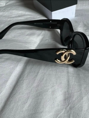 Svart Chanel solglasögon med guldlogo - Säljer ett par svarta solglasögon från Chanel med breda skalmar och en stor guldig Chanel-logo på sidorna. Glasögonen har en klassisk, chunky form och kommer med originalfodral. Perfekt accessoar för att lyfta vilken outfit som helst.