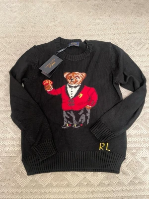 Svart stickad tröja Polo Ralph Lauren - Säljer en svart stickad tröja från Polo Ralph Lauren med ikoniska björnmotivet framtill. Björnen har röd kavaj och håller spelkort. Tröjan har ribbade muddar och rund hals. RL-broderi nertill. Perfekt för dig som vill ha något unikt och snyggt.