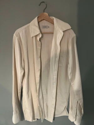 Vit manchester overshirt från SANDÖ - Säljer en stilren vit overshirt från SANDö i mjuk manchester. Skjortan har klassisk krage, långa ärmar och knappar framtill. Perfekt att bära öppen över en t-shirt eller stängd för en clean look. Enkel bröstficka och diskreta slitsar i sidorna. Aldrig använd!