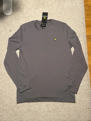 Grå långärmad t-shirt Lyle & Scott - Snygg grå långärmad t-shirt från Lyle & Scott Junior med klassisk rund hals och ikonisk gul logga på bröstet. Tillverkad i mjuk bomull som känns skön mot huden. Perfekt till chill vardagsstil och enkel att matcha med jeans eller shorts.