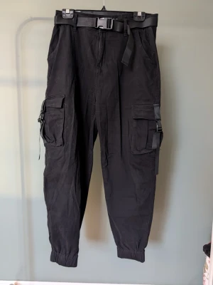 Svarta cargopants med bälte - Säljer ett par svarta cargopants med elastiska benslut och stora fickor på sidorna. Byxorna har ett justerbart bälte med plastspänne i midjan och coola detaljer med remmar på fickorna. Perfekta för en streetwear-look och riktigt sköna att bära.