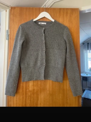 Grå stickad kofta från Zara - Snygg grå stickad kofta från Zara med rund hals och silverfärgade knappar framtill. Koftan har lång ärm och är i mjukt material som känns skönt mot huden..
