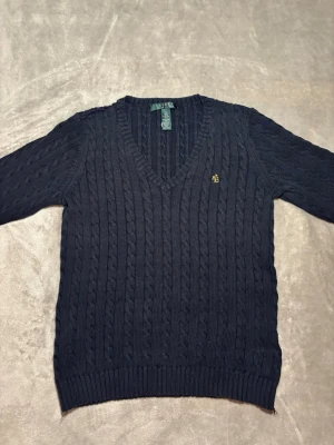 Svart kabelstickad tröja Ralph Lauren - Kabelstickad Svaft tröja från Ralph Lauren med v-ringning och broderad gul RL-logga på bröstet. Tröjan har lång ärm, ribbade muddar och är gjord i mjuk bomull. Perfekt för lager-på-lager under höst och vinter.