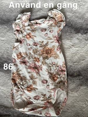 Blommig body från Newbie stl 86 - Supersöt blommig body från Newbie. Ljus botten med rosa och beige blommor, volangdetaljer på axlarna och spetskant. Perfekt för vår och sommar, lätt att knäppa undertill.🌸 Använd en gång. Storlek 86