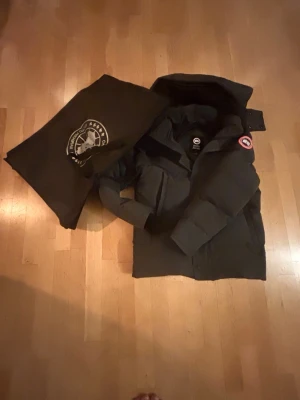 Grå dunjacka från Canada Goose WYNDHAM - Säljer en grå dunjacka från Canada Goose med avtagbar huva och pälsdetalj. Jackan har klassisk logga på ärmen, stora fickor och stängs med både dragkedja och knappar. Perfekt för kalla vinterdagar och riktigt snygg streetstil.