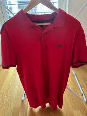 Röd slim fit piké från Hugo Boss - Säljer en röd pikétröja från Hugo Boss i storlek L. Modellen är slim fit och har klassisk krage, knappar framtill och diskret Boss-logga på bröstet. Ärmarna har svarta och röda ränder vid muddarna för en sportig touch.