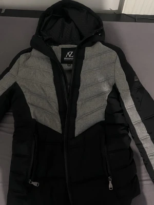 Svart och grå pufferjacka Alessandro Zavetti - Säljer en snygg svart och grå pufferjacka från Alessandro Zavetti i storlek XS. Jackan har huva, dragkedja framtill och två fickor med dragkedja. Fodret är mönstrat med logga och jackan har en modern, sportig look med paneler i grått och svart.